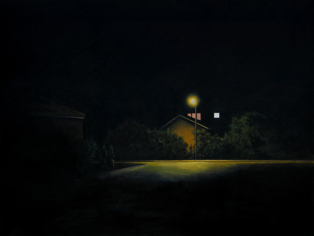 A moody nighttime scene dominated by a single, bright yellow streetlight. The light illuminates a patch of asphalt and the side of a nearby house nestled behind dark green bushes. In the background, two small windows—one pinkish-red and one white—provide a